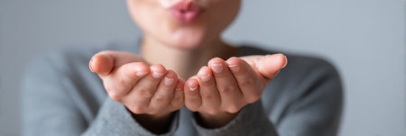 Hands extend outward in a blowing kiss gesture, illuminated by soft warm light with a simple, neutral background creating a serene atmosphere.の素材