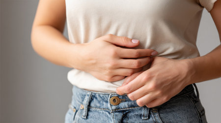 Woman is holding her belly fat with one hand while wearing a fitted shirt and denim jeans in a simple indoor setting.の素材