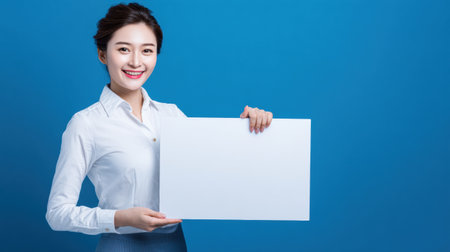 Smiling Asian woman stands in front of a bright blue background, holding a blank sign, ready to convey a message or information.の素材