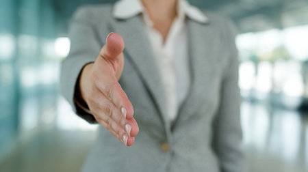 Businesswoman stands in an office and extends a handshake to a potential client. The setting is bright and professional during the afternoon.の素材