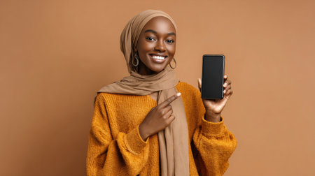 A joyful Muslim woman is smiling and pointing at her smartphone while dressed in a cozy outfit, showing her enthusiasm in a studio space.の素材