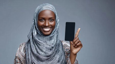 Cheerful muslim woman smiles and points at black smartphone screen in a simple, modern indoor environment, showcasing joy and engagement.の素材
