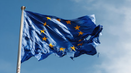 The European Union flag flutters steadily in the wind under a bright blue sky, showing its vibrant colors and iconic stars.の素材