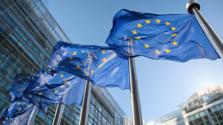 Multiple blue European Union flags wave on flagpoles outside the modern European Parliament building in Brussels under a sunny sky.の素材