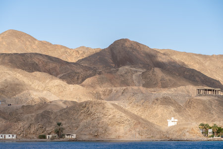 Rocky mountains rise prominently near a coastal area, with clear skies and sunlight casting shadows...