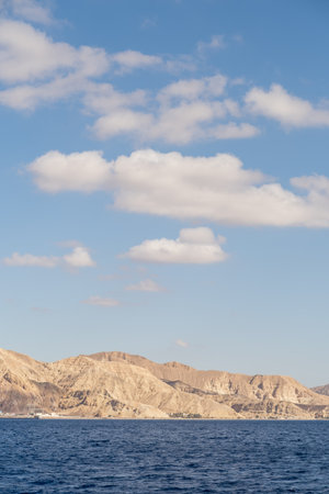 Coastal mountains rise majestically against a backdrop of blue sky and scattered clouds, creating a serene atmosphere over the calm sea.の写真素材