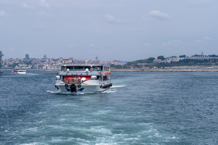 A ferry glides along the Bosphorus, creating ripples in the clear water, while the lively city landscape unfolds behind it under a bright skyの写真素材