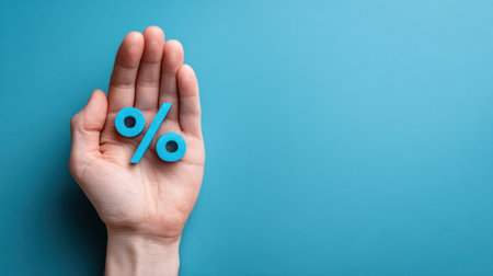 A hand is holding a blue percent symbol against a blank blue background, representing concepts in finance, discounts, and rates in a business setting.の素材