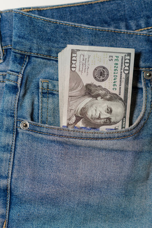 A stack of cash is partially visible in the front pocket of blue denim jeans, highlighting a casual...