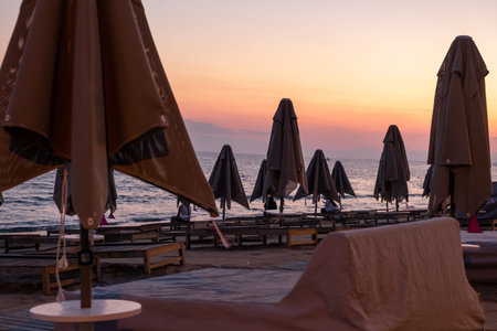 Relaxing beach setting features neatly arranged umbrellas and loungers as the sun sets, casting warm hues over the tranquil water and picturesque horizon.の写真素材