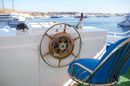 Boat steering wheel and blue chair positioned on yacht deck, with a marina and multiple boats visible in the background under a clear blue skyの写真素材