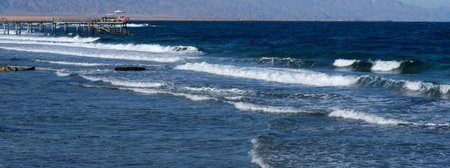 Coastal scene featuring waves breaking on the sandy beach, a wooden pier stretching into the ocean, and mountains visible in the distanceの写真素材
