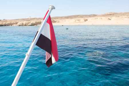 Egyptian flag displayed on a boat with vibrant blue waters and a sandy coastline visible in the background under a clear skyの写真素材