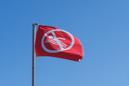 Red flag featuring a swimming prohibition symbol flutters in the wind against a bright blue sky, signaling no swimming in the vicinityの写真素材