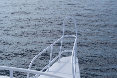White railing on a boat's bow extends over calm blue water, with gentle waves reflecting sunlight under a clear skyの写真素材