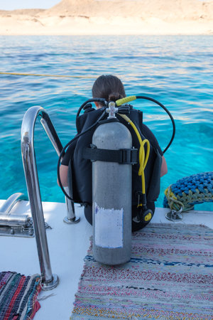 Diver sits on the edge of a boat, equipped with a scuba tank and life vest, ready to enter the clear...