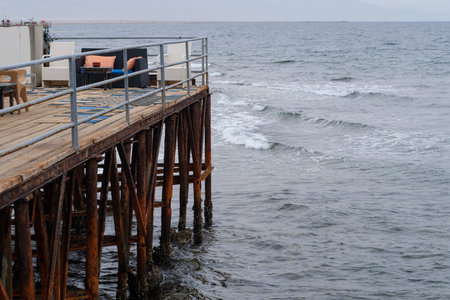 Wooden pier stretches over tranquil water with gentle waves, showcasing a chair and table set for...