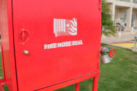 Red fire hose reel cabinet with a hose and valve, positioned against a wall in a grassy area, with a...