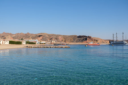 Coastal scene featuring a tranquil beach with clear water, rocky hills in the background, and boats...