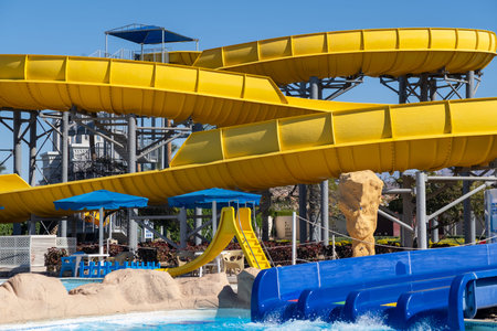 Large yellow water slides twist above a blue pool area in an amusement park, surrounded by sandy...