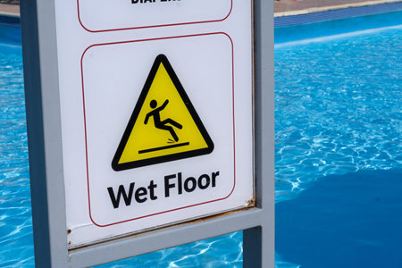 Caution sign warns of wet floor near swimming pool, featuring clear blue water and a tiled edge in...