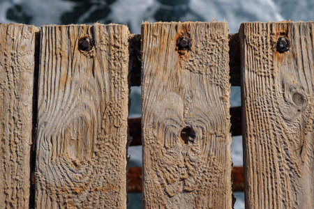 Close-up of aged wooden planks on a dock, featuring nails and a blurred water surface in the...