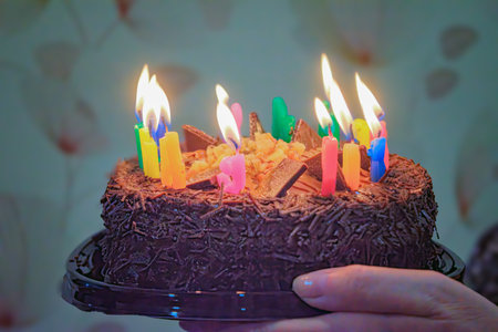 Woman holding birthday cake with burning candles against blurred background, closeup.