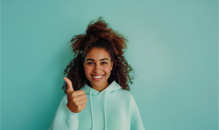 Young curly african american girl in a sweatshirt smiling with healthy teeth showing thumb up at copy space expressing wow emotion standing isolated on mint backgroundの素材