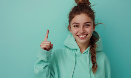 Smiling caucasian teenager girl in a mint sweatshirt smiling with healthy teeth and pointing index finger up at copy space expressing wow emotion standing isolated on paper mint backgroundの素材