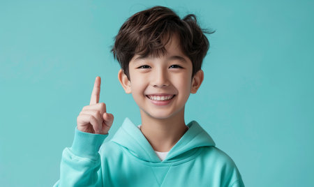 Smiling asian teenager boy in a mint sweatshirt smiling with healthy teeth and pointing index finger up at copy space expressing wow emotion on a mint backgroundの素材