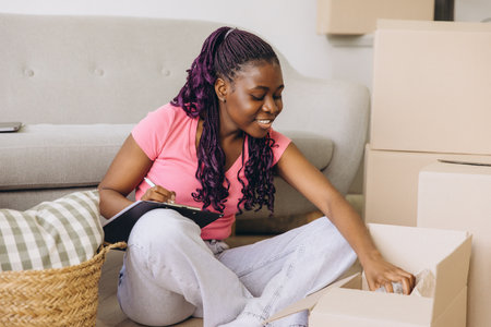 Happy African American woman taking notes on clipboard while unpacking cardboard boxes in new apartmentの写真素材