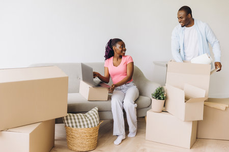 Young African American couple unpacking boxes and using laptop in new apartment, starting new life...