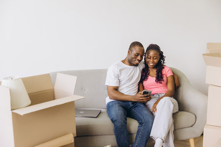 Happy African American couple enjoying a moment of relaxation on the sofa, using a smartphone during a break from moving into their new homeの写真素材