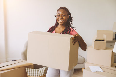 Smiling young african american woman carrying big cardboard box moving in new apartment surrounded...