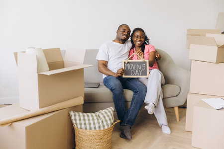 Happy African American couple showing moving day sign while sitting on sofa in new apartment surrounded by cardboard boxesの写真素材