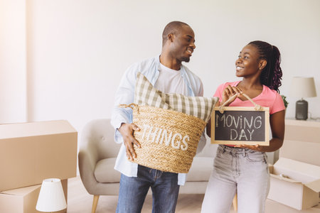 Smiling African American couple joyfully moving into their new apartment, holding a basket and a sign celebrating moving dayの写真素材