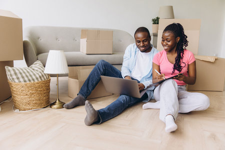 Happy African American couple taking inventory of their belongings while sitting on the floor in their new apartment, surrounded by cardboard boxesの写真素材