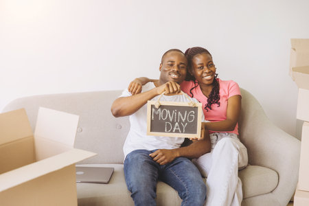 Happy African American couple sitting on a sofa in their new apartment, joyfully holding a moving day sign and smiling togetherの写真素材