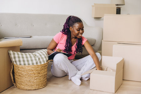 Happy African American woman unpacking boxes in a new apartment, organizing belongings, and jotting notes on a clipboardの写真素材