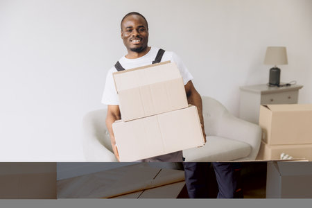 Smiling African American delivery man carrying cardboard boxes while moving into a new apartment, embracing the excitement of relocationの写真素材