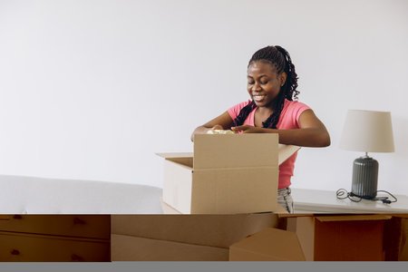 Smiling african american woman unpacking belongings from cardboard boxes in new apartment, starting new life after moving homeの写真素材