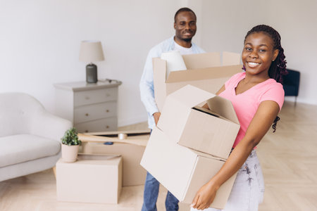 Young African American couple carrying cardboard boxes while moving into their new apartment, excitedly starting their life togetherの写真素材
