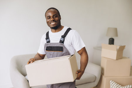 Young african american delivery man holding cardboard box while smiling and carrying belongings to new apartmentの写真素材
