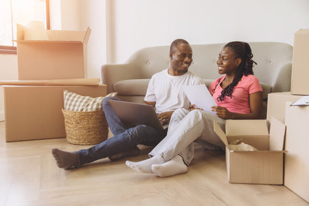 Happy african american couple sitting on the floor among cardboard boxes, using laptop and reading documents, organizing move in new apartmentの写真素材