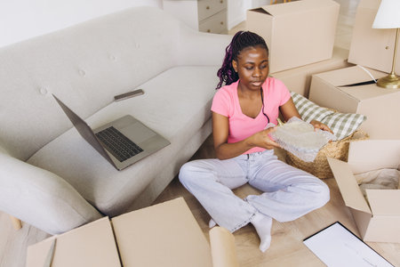 African American woman sitting on the floor, packing items into boxes for moving. A laptop and couch are nearby, indicating a home settingの写真素材