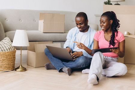 Happy african american couple sitting on floor among cardboard boxes using laptop for planning new home interior designの写真素材