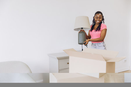 Happy young african american woman unpacking a lamp in her new apartment after moving house, surrounded by cardboard boxesの写真素材