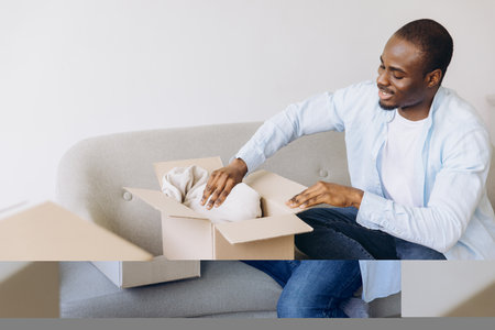 Happy African American man unpacking clothes from a cardboard box, settling into a new apartment and embracing the excitement of a fresh startの写真素材