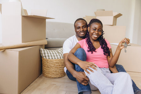 Young cheerful african american couple is hugging and showing keys of their new apartment while sitting among cardboard boxesの写真素材