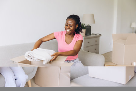 Smiling african american woman unpacking clothes from a cardboard box while sitting comfortably on a sofa in her bright, new apartmentの写真素材